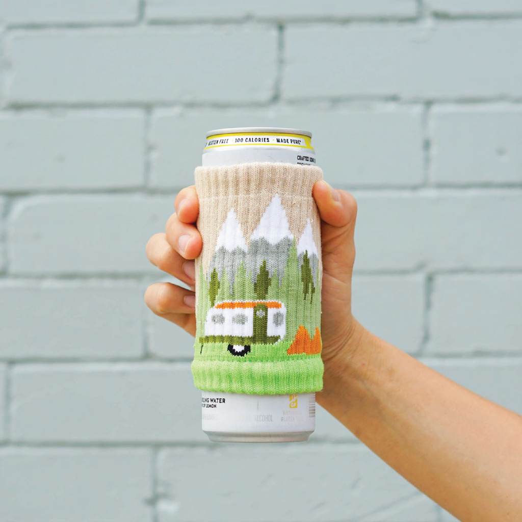 A seltzer can covered with a knitted drink sleeve that depicts a camper in the mountains